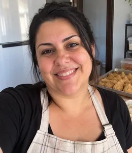 Criadora do método, de avental, na cozinha, com bandejas de salgados ao fundo